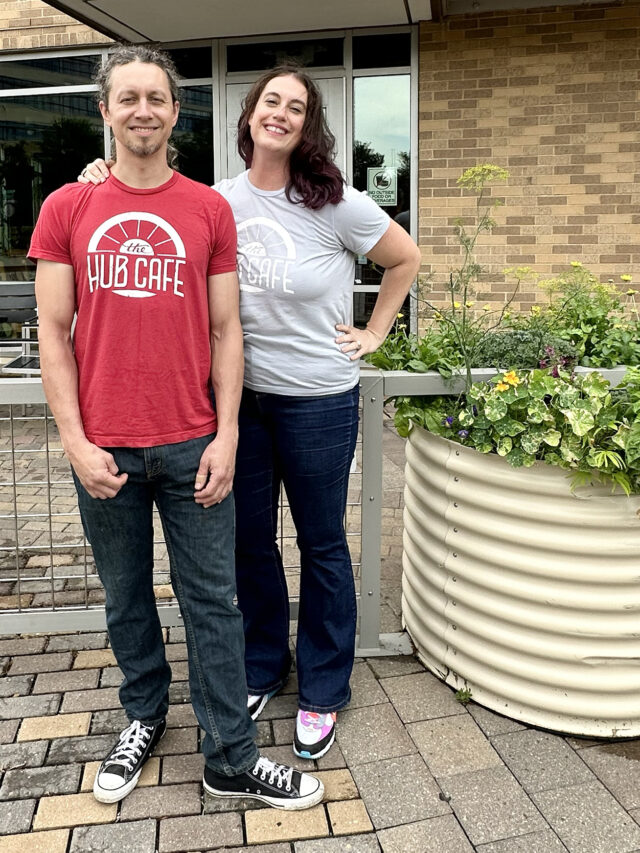 Couple who owns Hub Cafe restaurant standing in front of restaurant. Man on left in red shirt, woman on right in white shirt.